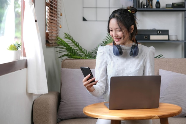 Feliz e alegre jovem asiática escolhendo sua lista de reprodução de música no celular ouvindo música através de seus fones de ouvido enquanto relaxa na sala de estar