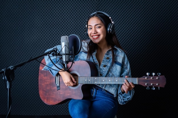 Feliz e alegre sorrindo de retrato de uma jovem vocalista asiática usando fones de ouvido com uma guitarra gravando uma música na frente do microfone em um estúdio profissional