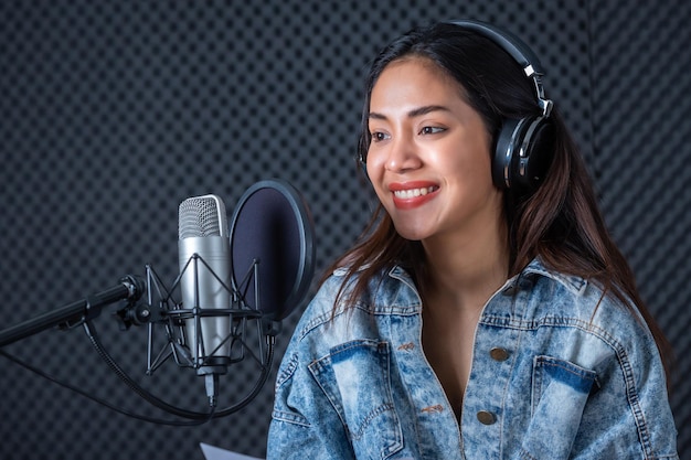 Feliz e alegre sorrindo do retrato de uma jovem vocalista asiática usando fones de ouvido gravando uma música na frente do microfone em um estúdio profissional