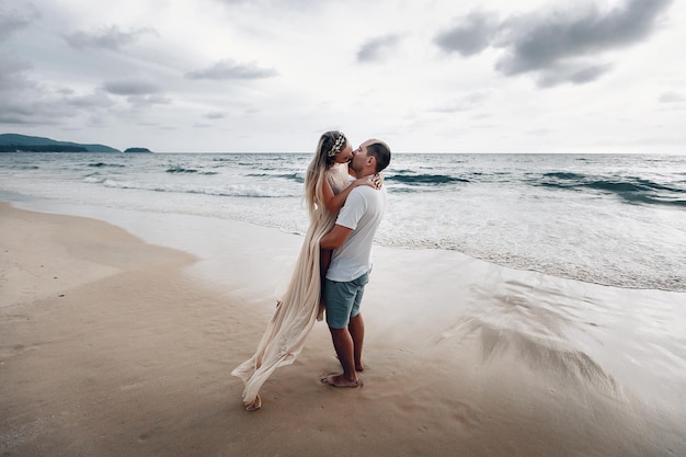 Feliz e amoroso marido e mulher vestidos de branco se beijando em uma praia exótica, o homem abraça a garota enquanto a levanta do chão.