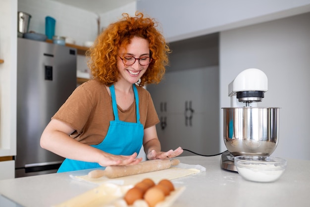 Feliz e atraente jovem adulta dona de casa padeiro usar avental segurando pino rolando massa na mesa da cozinha assando pastelaria conceito cozinhar bolo biscoito fazendo padaria fazendo pizza caseira em casa