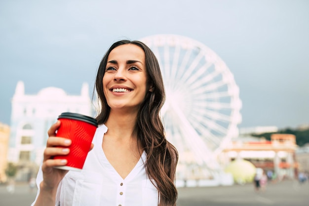 Feliz e bonita jovem sorridente está bebendo café enquanto caminha na cidade