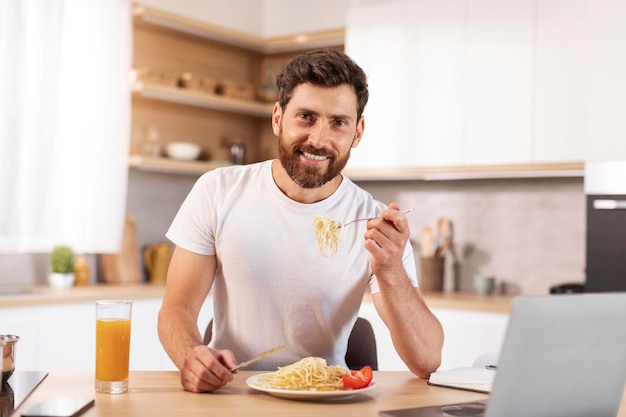 Feliz e bonito homem caucasiano barbudo comendo macarrão no interior da cozinha minimalista com laptop