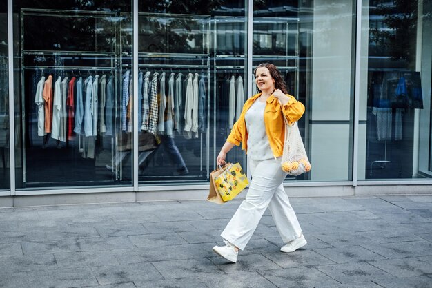 Feliz e confiante sorridente jovem curvilínea de tamanho grande com sacolas de compras e bolsa de rede da moda andando na rua da cidade perto de vitrines de shopping