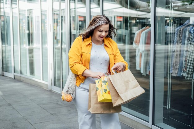 Feliz e confiante sorridente jovem curvilínea de tamanho grande com sacolas de compras e bolsa de rede da moda andando na rua da cidade perto de vitrines de shopping