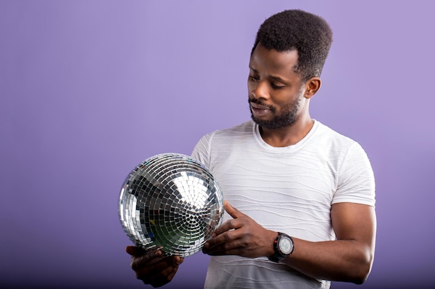 Feliz e encantador estudante de pele escura segurando uma bola de discoteca de prata brilhante, indo para a festa com amigos, celebrando a aprovação nos exames usando roupas casuais, sorrindo em pé sobre um fundo roxo.