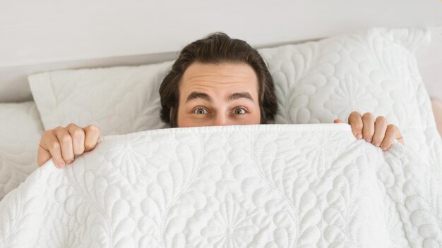 Foto feliz e engraçado homem europeu do milênio está deitado na cama branca coberta com cobertor, aproveite o tempo livre do fim de semana