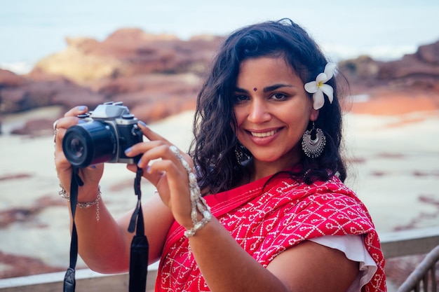 Feliz e linda mulher indiana no tradicional sari vermelho elegante segurando a câmera na mão posando para o fotógrafo na praia paraíso tropical mar oceano