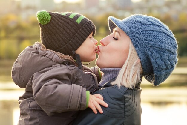 Feliz e linda mulher loira com chapéu, beijando o filho bebê com uma jaqueta quente, segurando-o nos braços no parque outono.