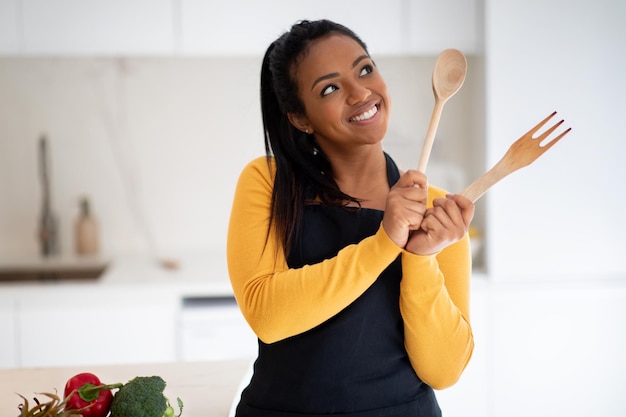 Feliz e pensativa jovem afro-americana cozinhando no avental perto da mesa com legumes pensa em sonhos com garfo de madeira