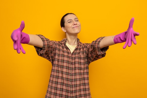 Feliz e positiva, jovem faxineira com camisa xadrez e luvas de borracha, dando boas-vindas ao geture, abrindo as mãos sorrindo em pé sobre a parede laranja