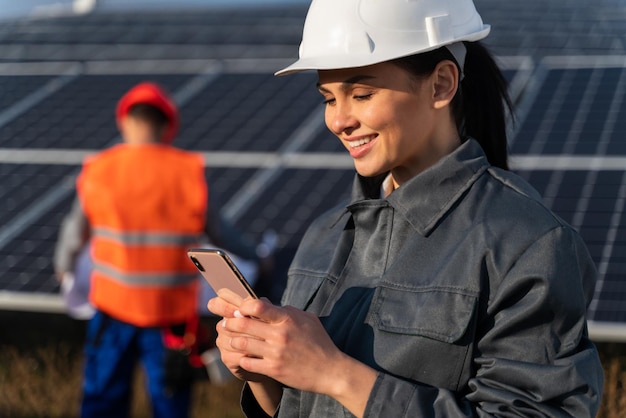 Feliz e sorridente engenheira no capacete está digitando algo no smartphone na bateria de energia no campo