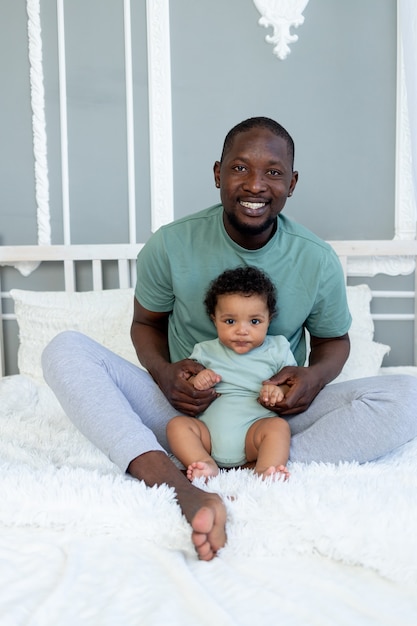 Feliz e sorridente pai afro-americano com o filho bebê na cama em casa, acariciando e brincando, família feliz, dia dos pais