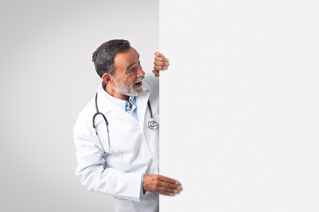 Feliz e surpreso velho médico de jaleco branco com estetoscópio olha para um grande banner com espaço vazio