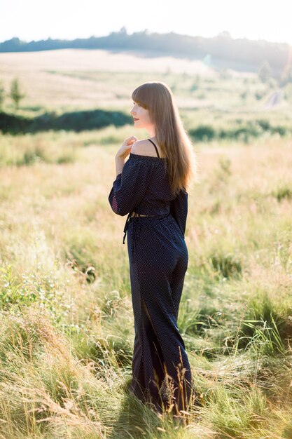 Feliz elegante jovem morena com roupas pretas, curtindo a natureza, posando em um lindo campo verde de verão ao pôr do sol