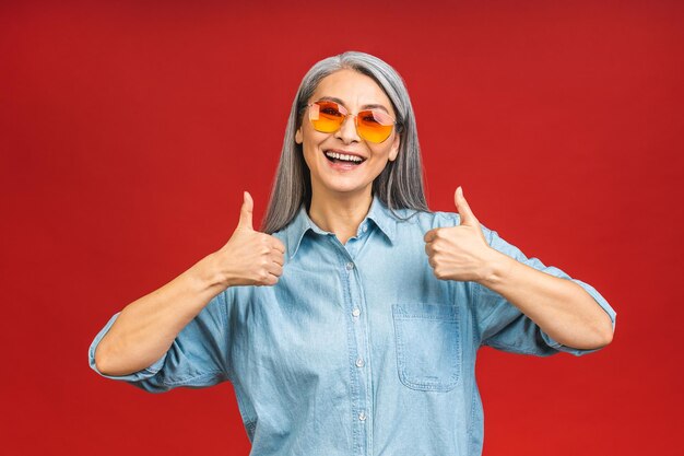 Foto feliz, elegante, madura, anciana, mujer de negocios asiática, sonriente, aislada, sobre fondo rojo, sonriente, confiada, alegre, de mediana edad, dama, espacio de copia, pulgares arriba, señal