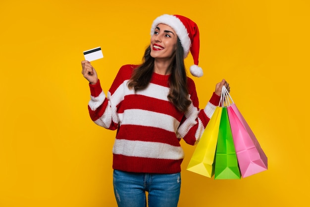 Feliz elegante mulher bonita com chapéu de Natal com sacolas de compras coloridas isoladas em fundo amarelo