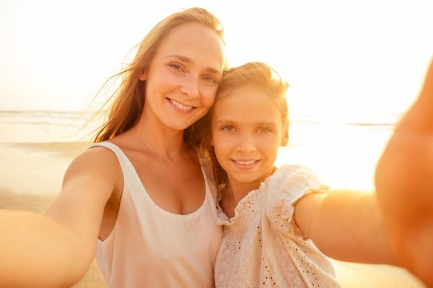 Feliz elegantemente mãe e filha tomando selfie na praia de areia em um pôr do sol, mãe, menina loira e linda mulher tirando fotos no telefone turismo no exterior chamadas de vídeo on-line