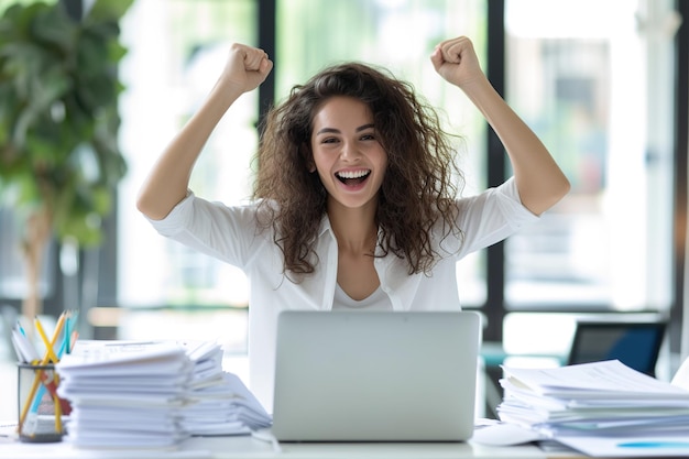 Foto feliz y emocionada joven mujer de negocios hispana contadora de pie en el escritorio trabajando en una computadora portátil con una pila de documentos en la mesa en la oficina y haciendo el gesto del sí regocijándose en un trabajo exitoso