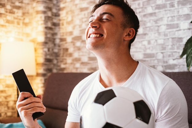 Feliz y emocionado hincha de fútbol viendo un partido en la televisión sosteniendo una pelota de fútbol en la mano y un control remoto mientras se sienta en un cómodo sofá en su sala de estar