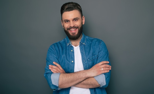 Feliz emocionado joven barbudo en jeans con los brazos cruzados está planteando sobre la superficie gris