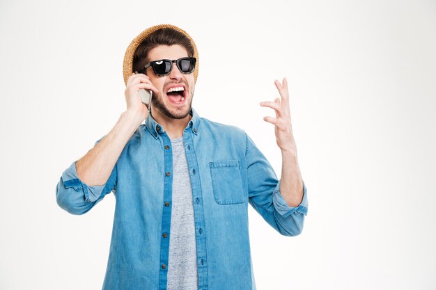 Feliz emocionado joven con sombrero y gafas de sol hablando por teléfono celular