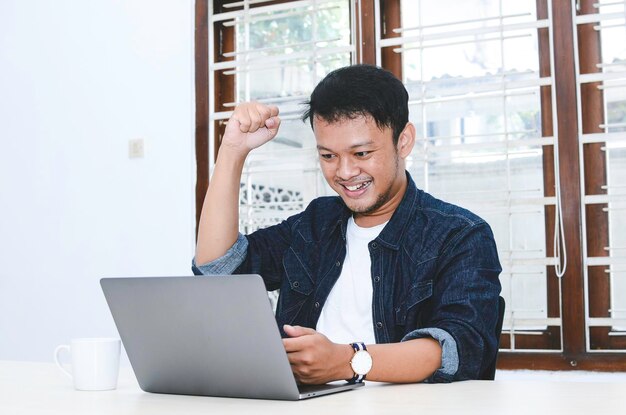 Feliz, emocionado y sonriente joven asiático levantando el brazo para celebrar el éxito o el logro cuando trabaja en una laptop Hombre indonesio con camisa azul