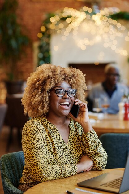 Feliz empresaria africana en anteojos riendo mientras tiene una conversación por teléfono móvil sentado en la mesa en el café