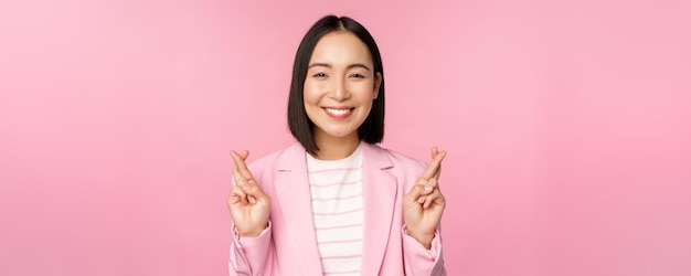 Feliz empresária asiática cruzar os dedos para boa sorte desejando orar e esperando sorrindo para a câmera em pé de terno sobre fundo rosa