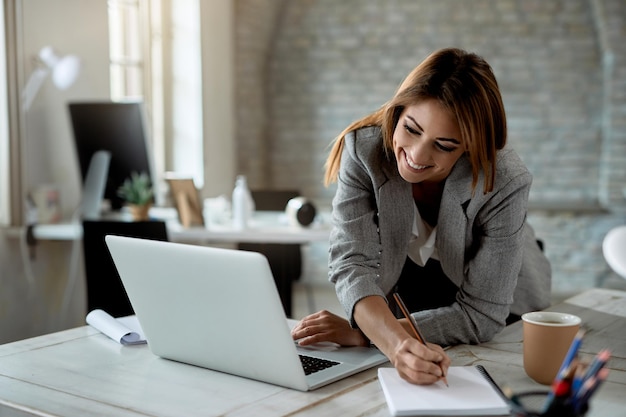 Feliz empresária escrevendo notas enquanto usa laptop e trabalha no escritório