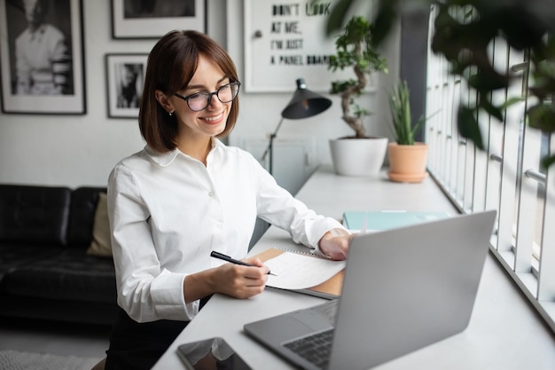 Feliz empresária europeia do milênio trabalhando em laptop fazendo anotações em espaço aconchegante de coworkign