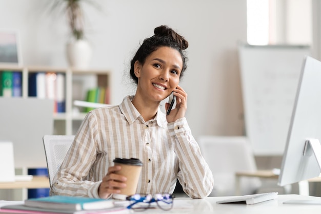 Foto feliz empresária falando no celular e tomando café no local de trabalho no escritório desfrutando de uma conversa agradável