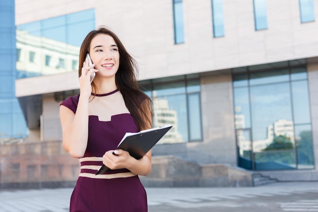 Feliz empresária falando no celular e trabalhando com documentos. Mulher trabalhando com papéis e consultoria em smartphone, perto do centro de escritório moderno, copie o espaço