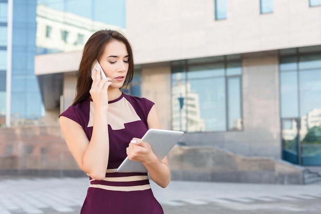 Feliz empresária falando no celular e trabalhando com tablet. Mulher segurando dispositivo digital e consultoria em smartphone, perto do centro de escritório moderno, copie o espaço