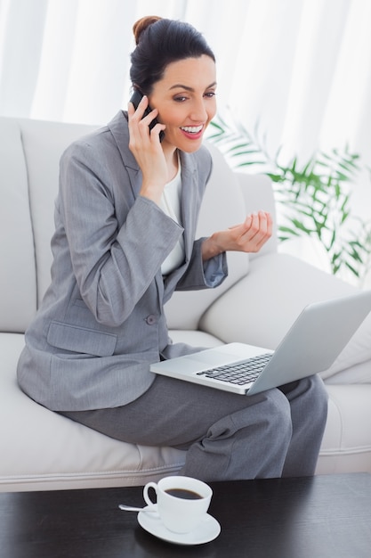 Feliz empresaria llamada con su teléfono móvil y el uso de la computadora portátil sentado en el sofá