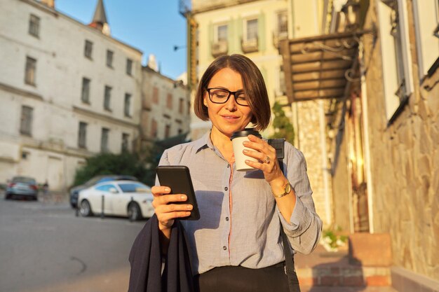 Feliz empresária madura em copos com café com smartphone na rua da cidade
