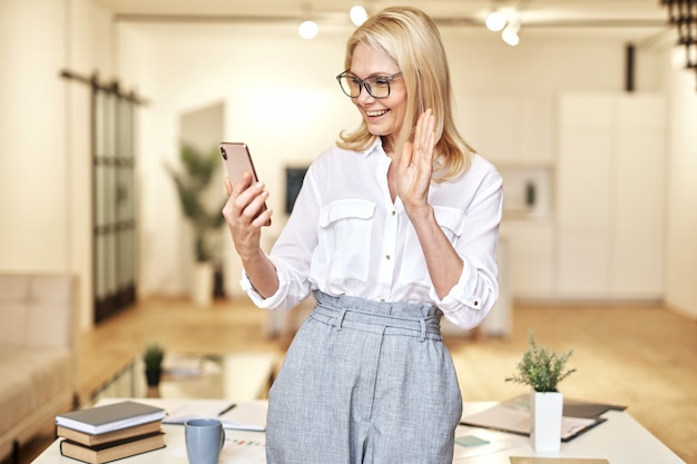 Feliz empresaria madura saludando a la cámara con videollamada en línea con smartphone mientras