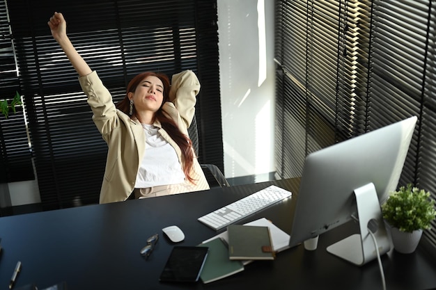 Feliz empresária milenar relaxada esticando os braços acima da cabeça em uma confortável cadeira de escritório