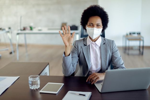 Feliz empresária negra com máscara facial trabalhando no escritório e acenando para a câmera