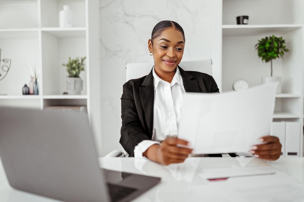 Feliz empresaria negra leyendo artículos comerciales y sonriendo comprobando las tasas de crecimiento de la empresa