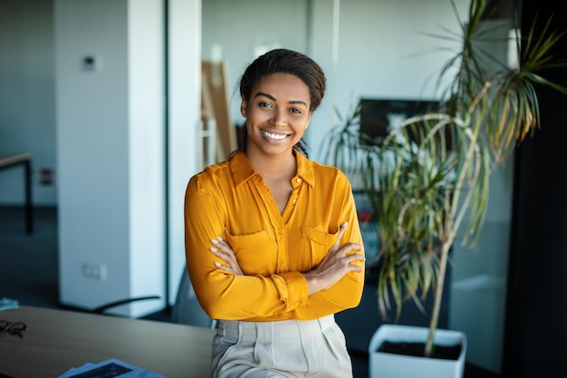 Feliz empresaria negra de pie con los brazos cruzados frente a su escritorio en la oficina moderna y sonriendo a la cámara