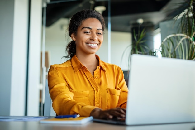 Foto feliz empresária negra usando laptop digitando e trabalhando online enquanto está sentado no local de trabalho no espaço de cópia do escritório