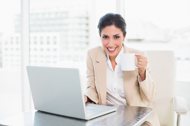 Feliz empresaria sosteniendo la taza mientras trabajaba en la computadora portátil