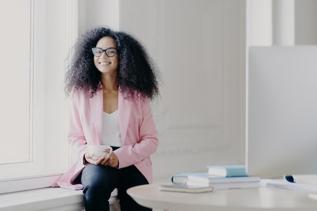 La feliz empresaria tiene el pelo afro tupido, usa una chaqueta rosada y pantalones negros, sostiene una taza de bebida en el alféizar de la ventana en un espacioso gabinete con una mesa, una computadora moderna disfruta de un café aromático