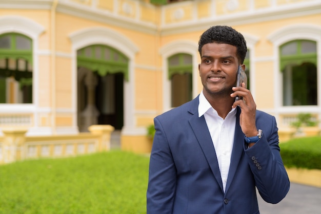 Feliz empresário africano ao ar livre, vestindo terno e falando no celular enquanto sorri, tiro horizontal