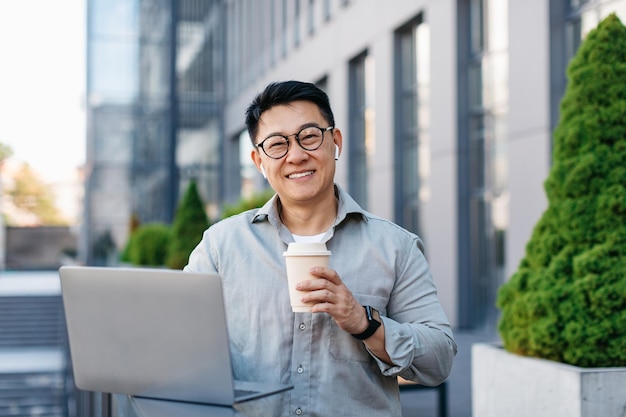 Feliz empresário asiático de meia idade trabalhando em laptop e bebendo café ao ar livre perto de prédio de escritórios