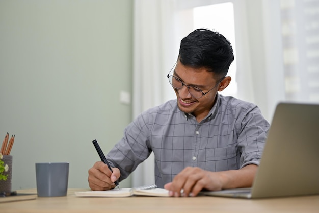 Feliz empresário asiático escrevendo algo em seu livro trabalhando em sua mesa no escritório