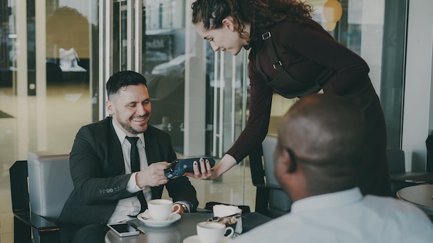Feliz empresário caucasiano barbudo sentado com seu colega afro-americano e pagando a conta on-line usando seu smartphone no café Garçonete sorridente usando terminal de dinheiro