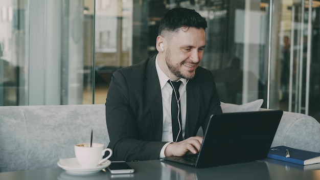 Feliz empresário caucasiano em roupas formais digitando em seu laptop enquanto ouve música com fones de ouvido em um café moderno durante o almoço