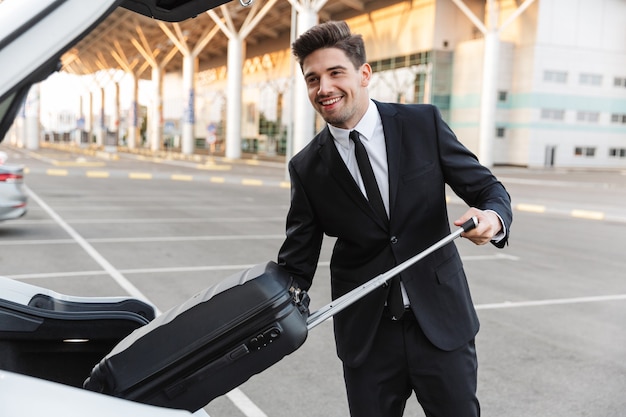 Feliz empresário com roupa formal colocando a mala no porta-malas do carro no estacionamento perto do aeroporto ao ar livre